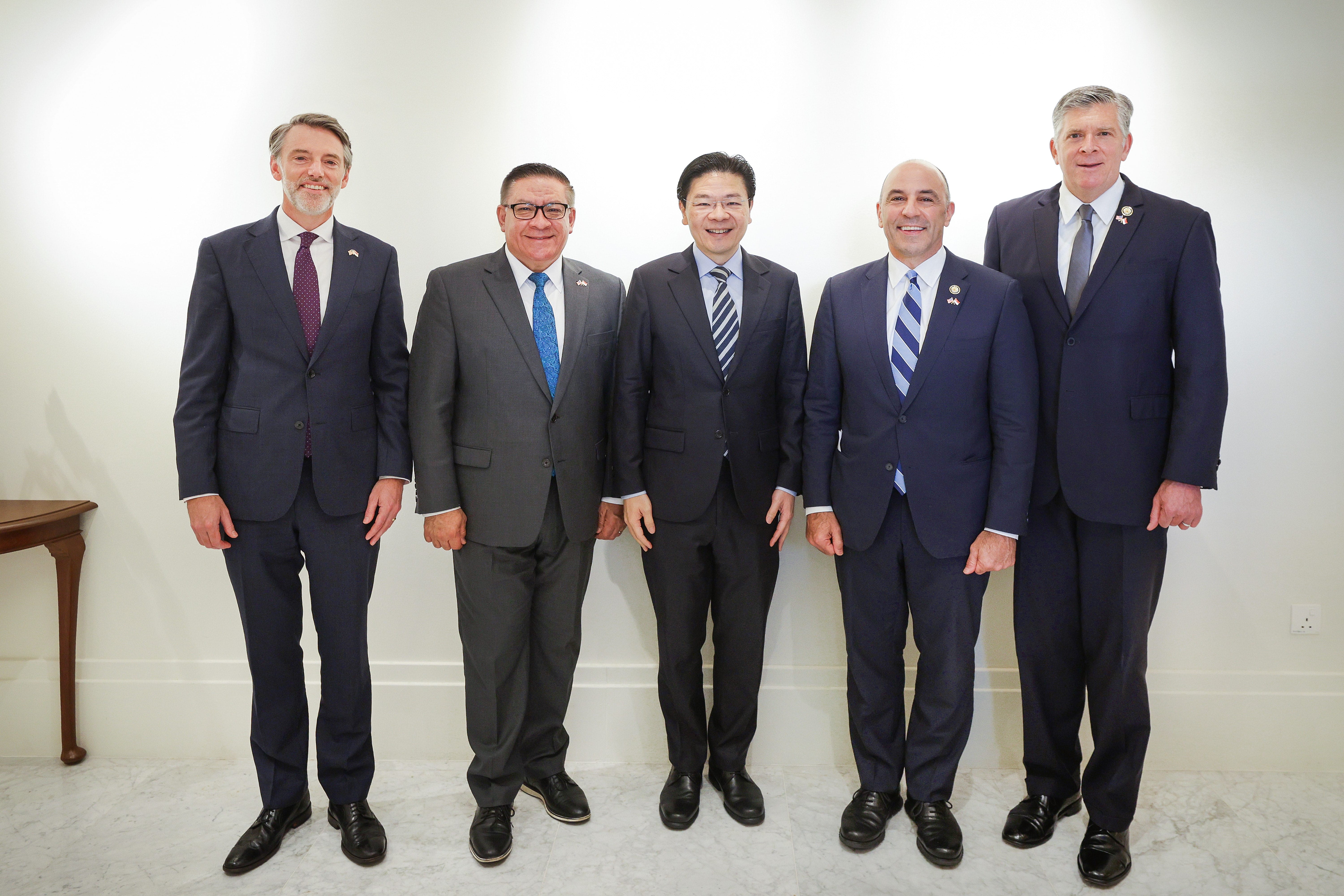 Five men in suits stand in a line against a white wall.